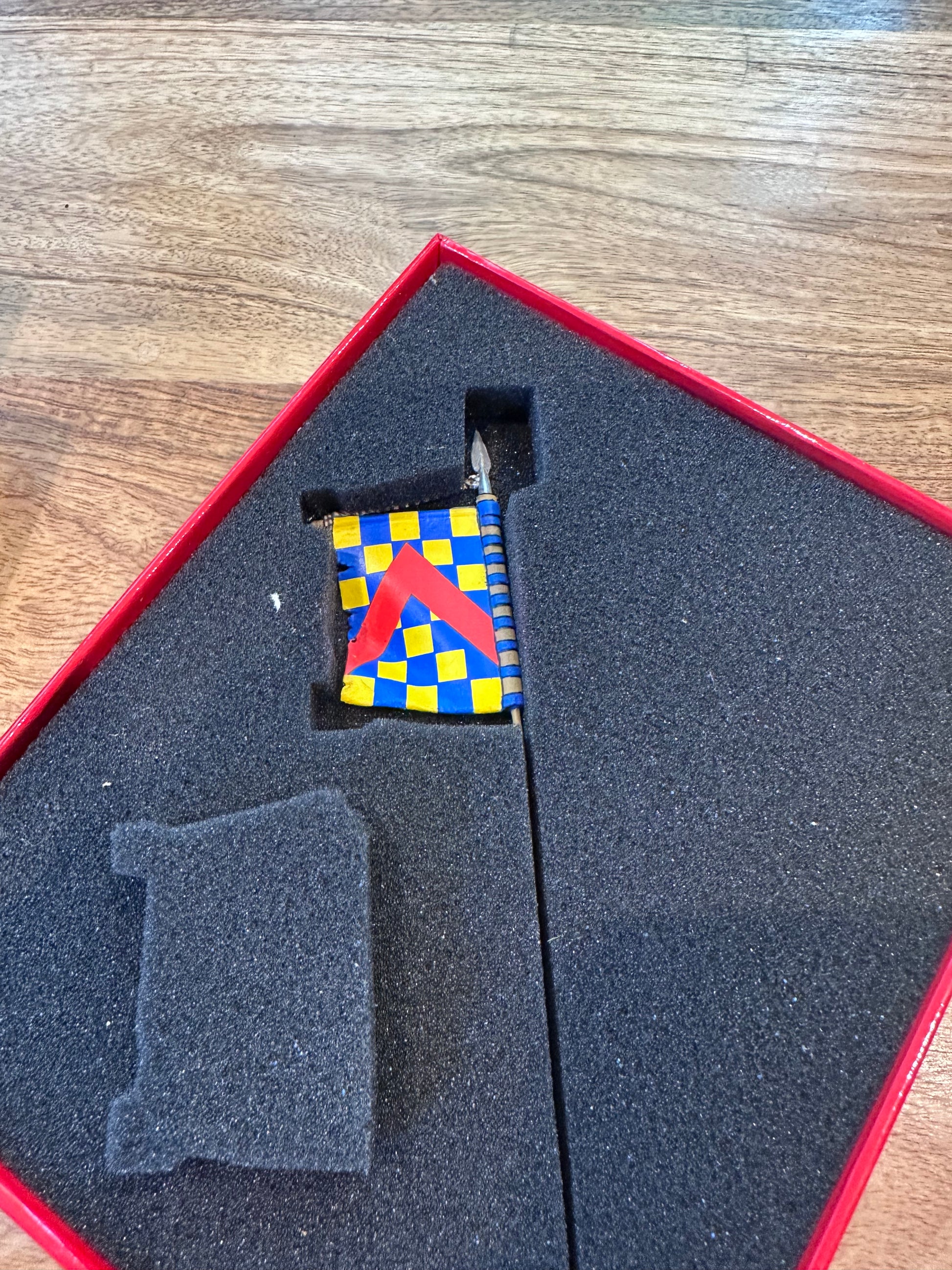 Small checkered flag in a red and black foam-lined box on a wooden surface for toy soldier Robert de Chevron.