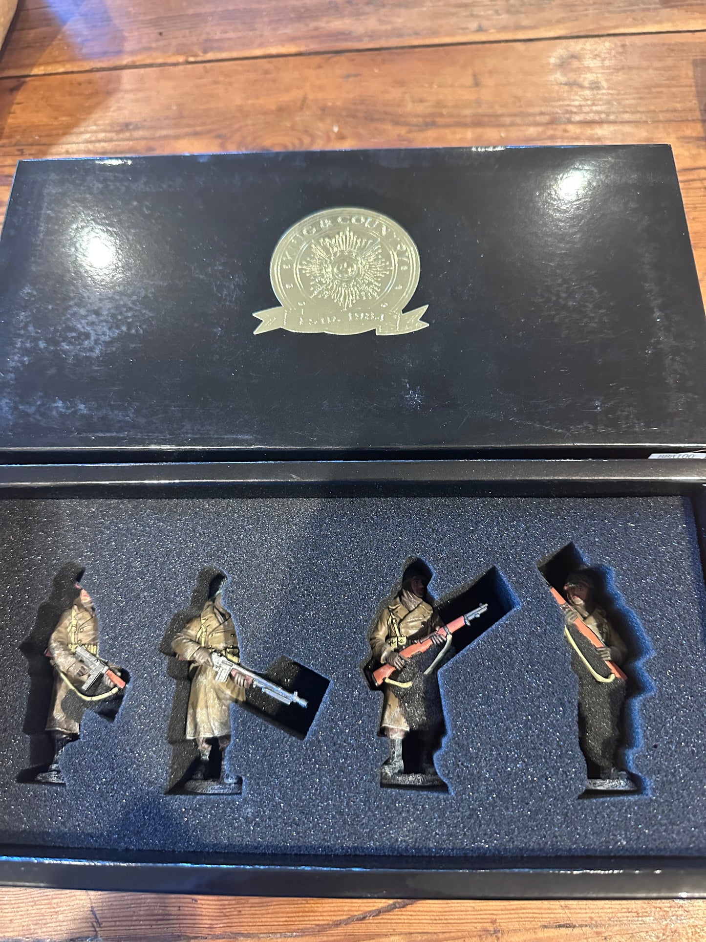 A set BBA100 of four miniature military figurines displayed inside an open black presentation box with a foam insert, placed on a wooden surface. The box lid features a metallic emblem.