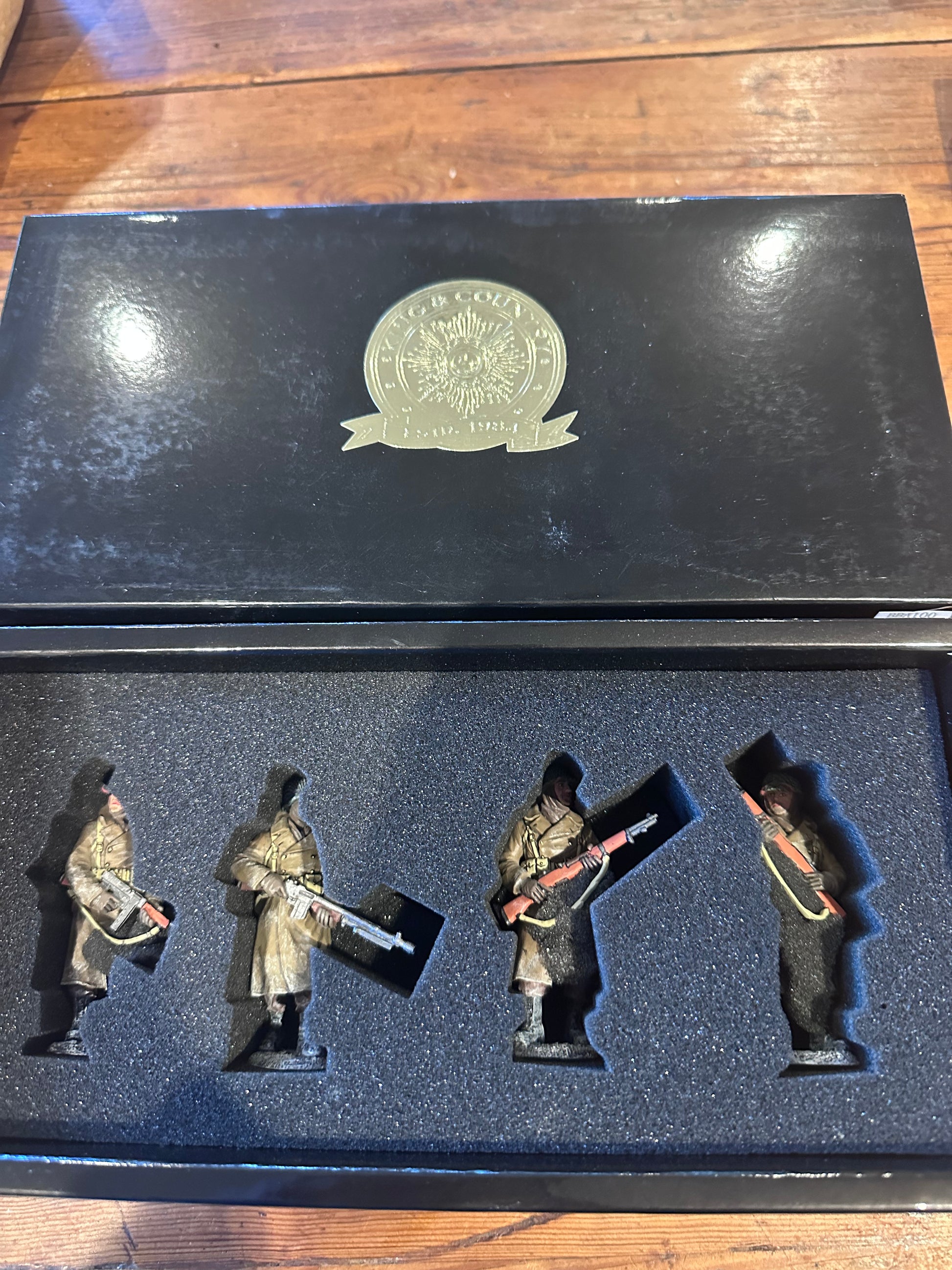 A set BBA100 of four miniature military figurines displayed inside an open black presentation box with a foam insert, placed on a wooden surface. The box lid features a metallic emblem.