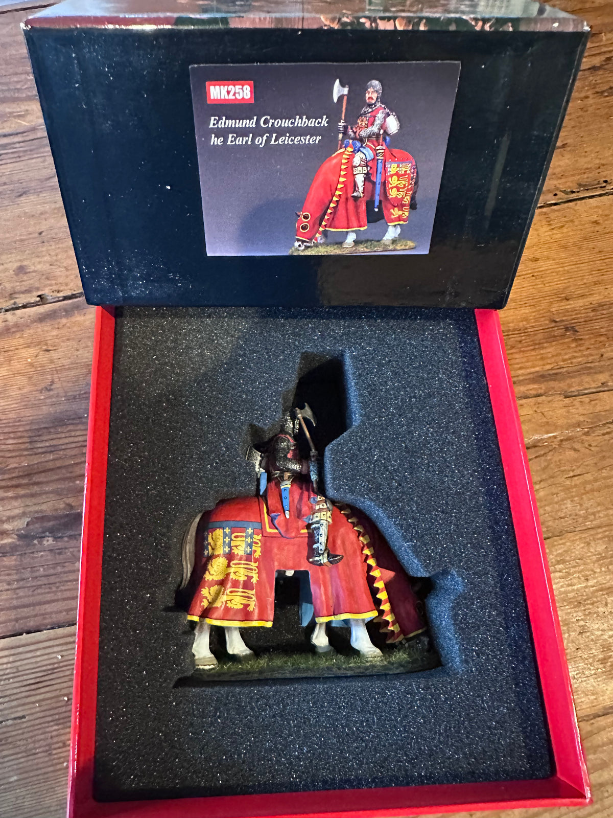 A boxed miniature figurine of a mounted soldier on horseback, shown open in its retail box on a wooden surface. The box interior is black foam with a red border, and the lid has a printed label reading 'MK258 Edmund Crouchback the Earl of Leicester.