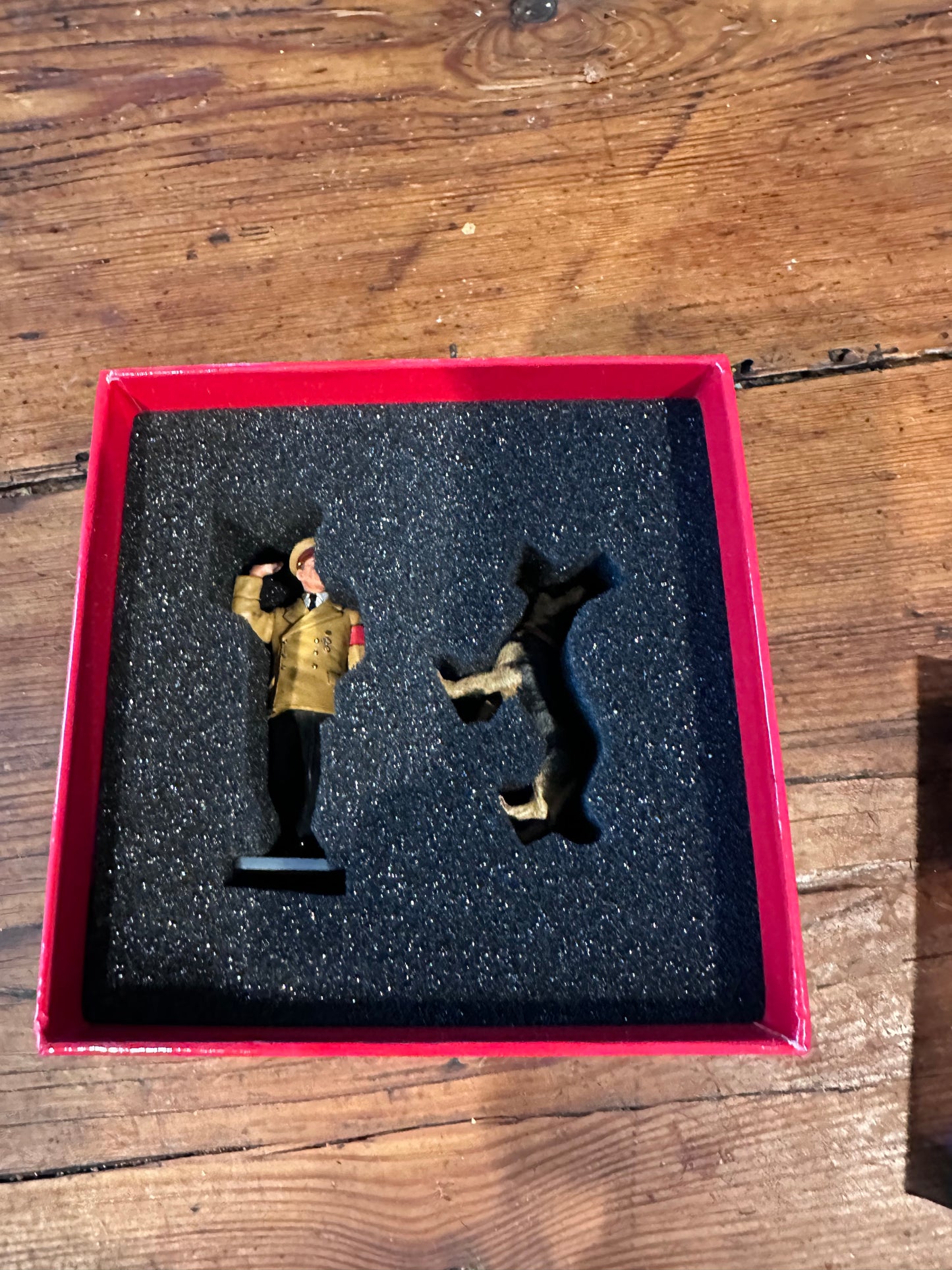 Two toy soldier Adolf and Blondi figurines on a dark foam insert in a red box on a wooden surface.