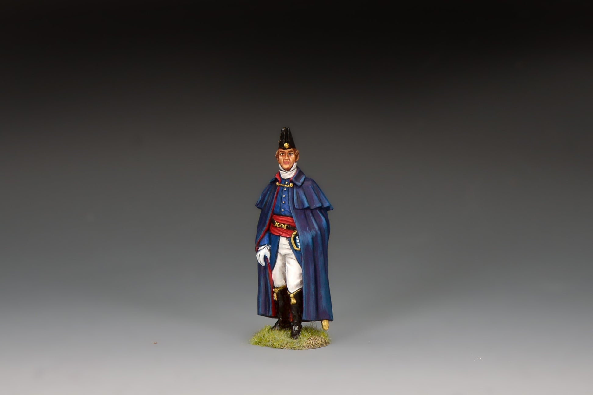 King & Country matte-painted metal figure of the Duke of Wellington standing in a dark blue caped cloak and black cocked hat, red sash, holding a riding crop, on a groundwork base.