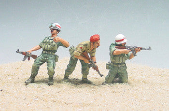 Three miniature toy soldiers in military uniforms stand on sandy ground. Two wear green outfits and white helmets, aiming rifles, while the center figure in tan uniform and red beret crouches, holding a weapon.