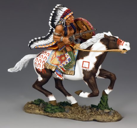 A painted toy soldier figurine of a Native American warrior in a feathered headdress riding a galloping brown and white horse, holding a rifle, with decorated clothing and horse accessories.