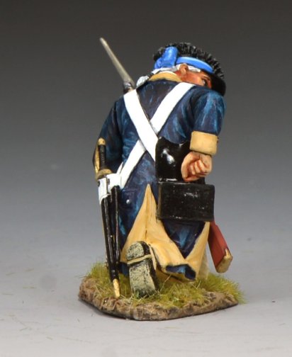 A toy soldier figurine of a soldier in colonial-era uniform kneels on one knee, holding a rifle with a bayonet. The soldier wears a blue coat, beige pants, and a blue cap, set against a neutral background.