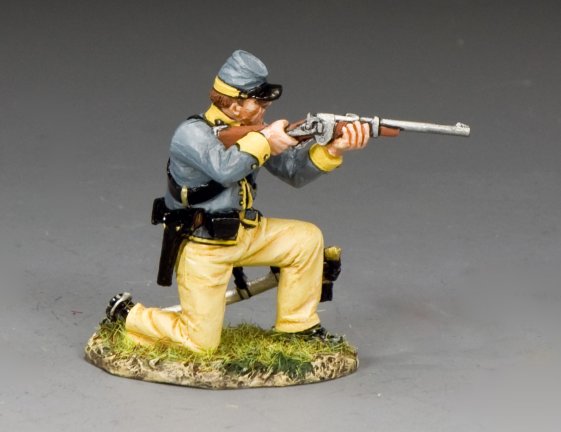 The King and Country Kneeling Trooper is a detailed miniature of a soldier in a blue-gray uniform with yellow pants, kneeling and aiming his rifle. He stands on a grassy base, wearing a cap, belt, and holster.