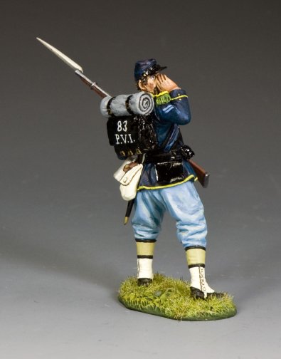 A toy soldier figurine in a blue uniform stands on grass, carrying a rifle and backpack labeled 83 P.V.I. The figure has a cap, light blue pants, and is cupping a hand to its mouth as if calling out.