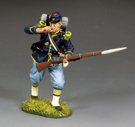 A detailed figurine of a Union Civil War soldier in blue uniform and cap, holding a rifle with a bayonet, stands on grass. The soldier raises one hand to his mouth as if shouting or calling out.