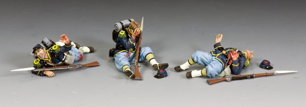 Three miniature toy soldiers in blue and light blue uniforms lay on a gray surface, appearing injured or resting, with backpacks and rifles beside them and hats on the ground.