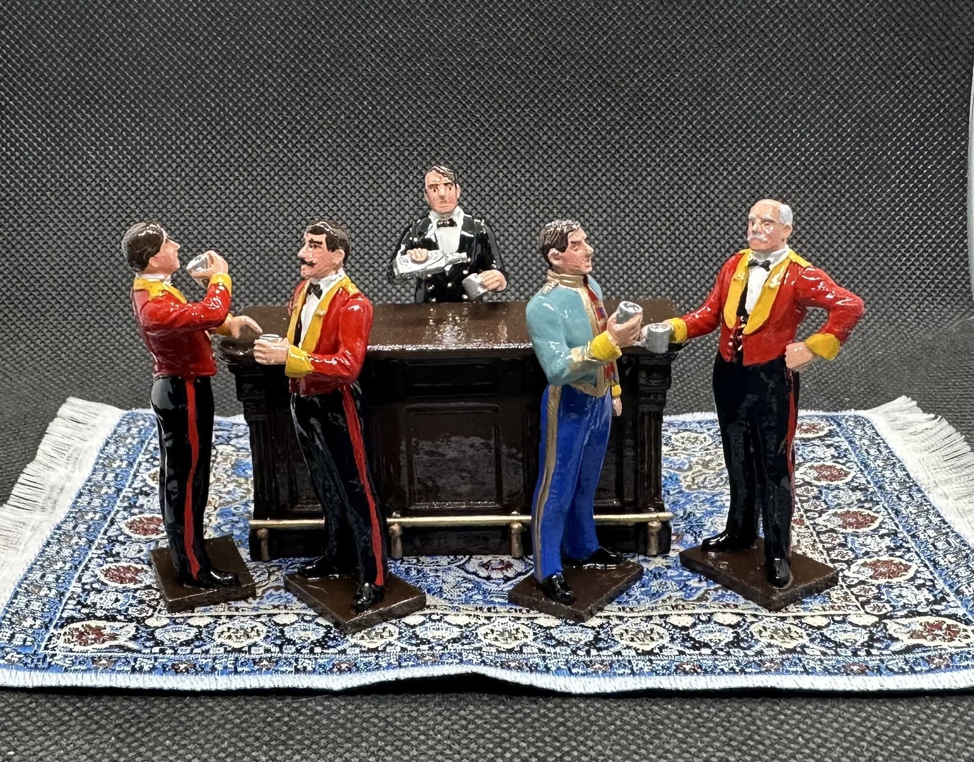 A set of five miniature figurines depicting men in waiter uniforms gathered around a bar counter, displayed on a patterned rug against a black mesh background.
