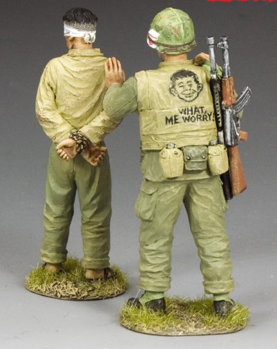 What Me Worry? Two toy soldiers in military uniforms with one holding a gun and the other with a blindfold, on a gray background.